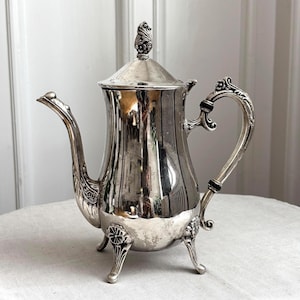 Puede incluir: Una tetera de tono plateado pulido con cuerpo cónico, pico curvo y un asa ornamentada con detalles en negro. La tapa tiene un remate decorativo y la base se apoya sobre tres patas adornadas con detalles en relieve de motivos florales.