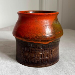 May include: A handcrafted ceramic vase with a unique design. The vase features a vibrant orange top that transitions to a dark brown base with a textured pattern. The interior is a dark brown color. A decorative piece for any home.