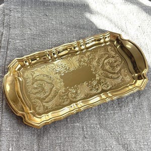 Op de afbeelding: Een rechthoekig goudkleurig metalen dienblad met afgeronde hoeken en geschulpte randen. Het oppervlak is voorzien van een ingewikkeld, in reliëf gemaakt krulpatroon rondom een glad, effen centraal gedeelte. Het dienblad heeft een gepolijste afwerking.