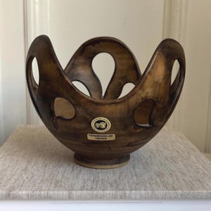 May include: A brown ceramic bowl with a distinctive, open design. The bowl features a rounded base and a series of arched openings along the rim. A small label with text is attached to the side. The bowl is likely handmade and suitable for decorative use.