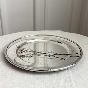 May include: A round, polished silver-toned metal serving platter featuring a smooth, reflective surface and a decorative rim with subtle, repeating cross-hatch detailing. A delicate, dried sprig of small pink and white flowers rests centrally.