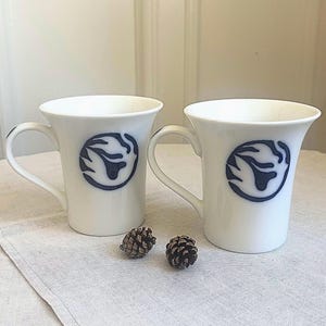 May include: Two white ceramic mugs with flared rims and curved handles. Each mug features a circular dark blue graphic design on the front. The design depicts a stylized abstract shape resembling a flame or a bird within a solid blue circle.