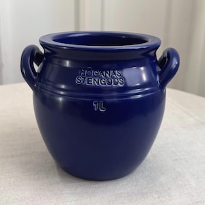 May include: A deep blue ceramic pot with two side handles and a wide opening. The front features the embossed text HOGANAS STENGODS and the capacity marking 1L. The vessel has a smooth, glossy finish and a rounded, bulbous body shape.