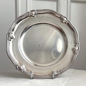 Peut inclure: Une assiette ronde en argent avec un bord festonné et des détails en relief décoratifs le long du rebord. La surface présente une inscription gravée "På 25 årsdagen 19-7-38" près du bord supérieur, avec un centre lisse et réfléchissant.