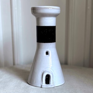 May include: A white ceramic lighthouse-shaped vase with a black band around the middle. The vase has a small door and window detail. The top rim has a thin gold accent. This decorative vase is suitable for home decor.