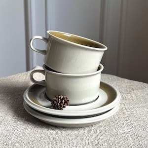 May include: Two stacked ceramic cups with matching saucers in a light cream color. The interior of the top cup and the rims of the saucers feature a muted olive-brown band. A small brown pinecone rests on the edge of the bottom cup's saucer.