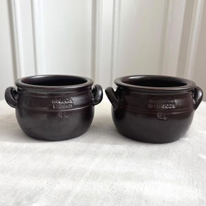 Op de afbeelding: Twee donkerbruine keramische potten met elk twee zijhandvatten. De voorzijde van elke pot is voorzien van de in reliëf aangebrachte tekst "HÖGANÄS STENGODS" en "1/2 L". De potten hebben een ronde vorm, een wijde opening en een glad glazuur.