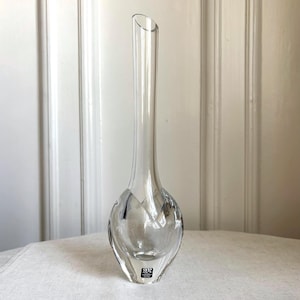 May include: A clear glass bud vase with a slender, elongated neck and a rounded, bulbous base. The glass features a faceted, geometric design around the lower section and includes a small black rectangular label with white text at the bottom.
