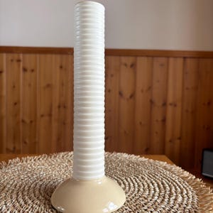 May include: A tall, cream-colored vase with a cylindrical, ribbed body and a wide, rounded base. The vase is approximately 12 inches tall, and sits on a woven placemat. The background features wooden paneling.
