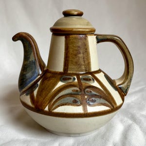 Può includere: Una teiera in ceramica con coperchio, beccuccio e manico. La teiera è color crema con motivi decorativi marroni e blu. Il coperchio e il manico hanno un bordo marrone. Il beccuccio ha un accento blu.
