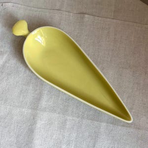 Könnte beinhalten: Eine gelbe Keramik-Servierschale in Tropfenform mit kleinem Griff. Die Schale hat einen weißen Rand und steht vor einem neutralen Hintergrund. Dieses dekorative Stück eignet sich zum Servieren von Vorspeisen oder kleinen Portionen.