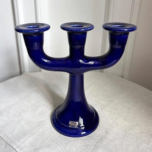 May include: A deep cobalt blue ceramic candelabra featuring three candle holders arranged in a symmetrical arc. The piece has a smooth, glossy finish and a wide, stable circular base. A small rectangular label is visible on the front of the base.
