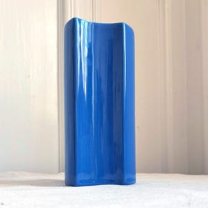 Könnte beinhalten: Eine hohe, zylindrische Keramikvase mit einer leuchtend blauen, glänzenden Oberfläche. Die Außenseite weist ein ausgeprägtes vertikales Wellenmuster auf, das tiefe Schatten und Lichtreflexe entlang des gesamten Objekts erzeugt.