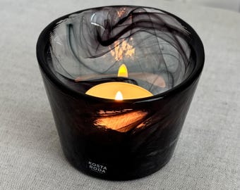 May include: A Kosta Boda glass votive candle holder featuring a dark, swirling black and translucent smoke-coloured pattern. The bowl-shaped vessel contains a lit tea light candle with a warm yellow flame, casting a soft glow through the glass.
