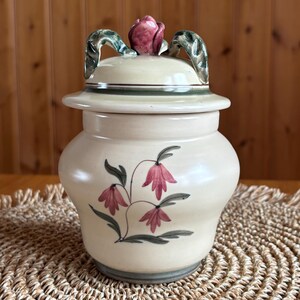 Könnte beinhalten: Ein cremefarbenes Keramikgefäß mit Deckel und floralem Design. Der Deckel hat einen knauf in Form einer rosa Rose und grüne, blattförmige Griffe. Das Gefäß ist mit rosa glockenförmigen Blüten und grünen Blättern verziert.