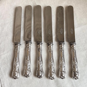 Könnte beinhalten: Ein Set aus sechs silberfarbenen Metallmessern, die in einer Reihe angeordnet sind. Jedes Messer hat eine glatte, flache Klinge und einen verzierten Griff mit geprägten Schnörkeln und muschelartigen Motiven am Ende. Polierte Oberfläche.