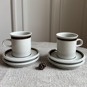 May include: Two ceramic cups and matching saucers in a light gray finish, each featuring a prominent dark brown horizontal band near the rim. The set is arranged with two stacked saucers under each cup, showcasing a simple, cylindrical design.