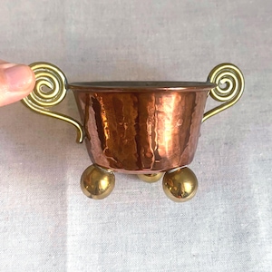 May include: A small, hammered copper bowl featuring a wide, tapered body with a metallic sheen. It is equipped with two decorative brass handles shaped like spirals and stands on three polished brass ball feet at the base of the vessel.