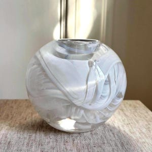 May include: A spherical glass vase featuring a clear base and a swirling white opaque pattern throughout the body. The vase has a circular opening at the top with a smooth, thick rim, showcasing a translucent quality and a rounded, bulbous shape.