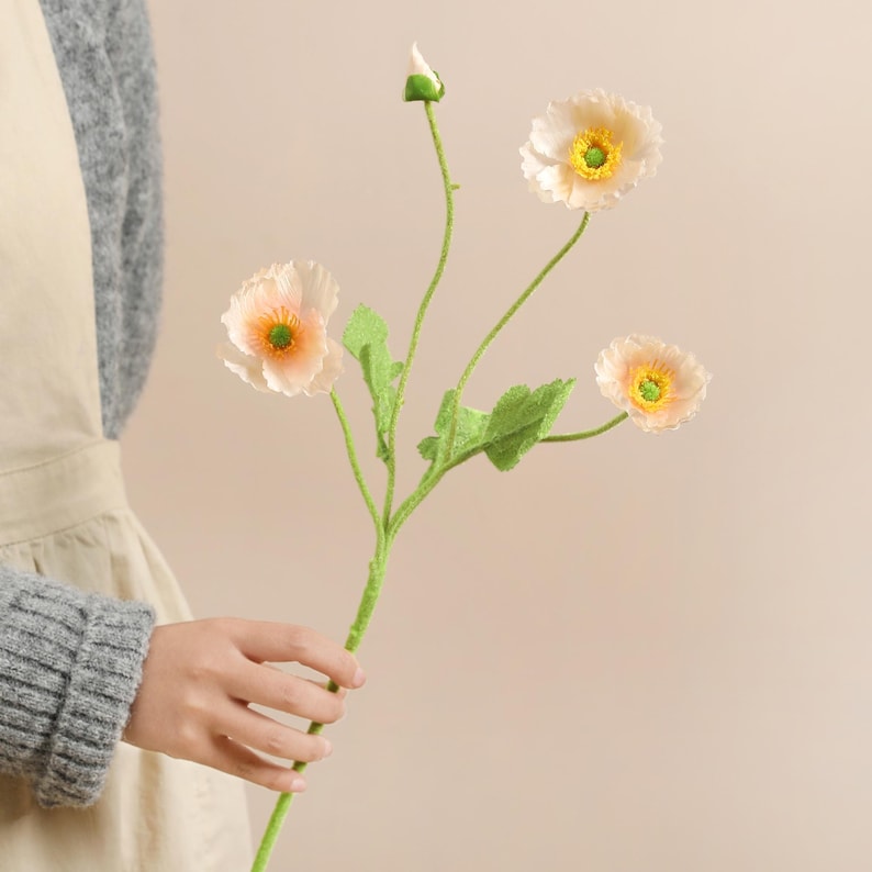 May include: A sprig of artificial poppies with four blooms and a bud. The flowers have soft peach-coloured petals with yellow and green centres. The stem is green with green leaves.