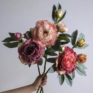 May include: A bouquet of artificial flowers featuring large, multi-layered peony-style blooms in shades of deep magenta, soft pink, and cream. The stems include several smaller, closed flower buds and numerous matte green leaves on thin stalks.
