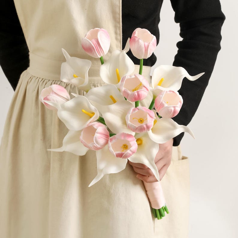 May include: A bouquet of white calla lilies and pink tulips. The flowers are held together with a pink ribbon. The calla lilies have yellow centers. The tulips have pink and white petals.