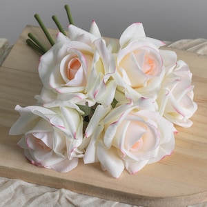 Könnte beinhalten: Ein Strauß weißer Rosen mit rosa umrandeten Blütenblättern und pfirsichfarbenen Zentren. Die künstlichen Blumen haben grüne Stiele und sind auf einer Holzoberfläche angeordnet, wodurch eine weiche und elegante Präsentation entsteht.