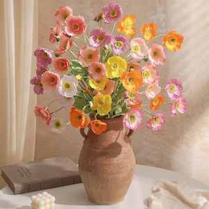 Op de afbeelding: Een terracotta vaas gevuld met kleurrijke kunstmatige klaprozen in roze, oranje, geel en wit. De bloemen hebben groene stelen en zijn gerangschikt in een vol boeket. De vaas staat op een witte tafel, met een boek en een kaars in de buurt.