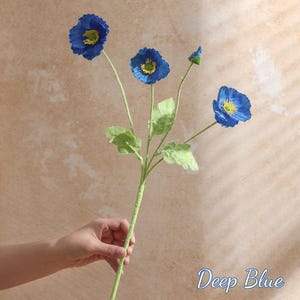 May include: A deep blue artificial poppy stem with four flowers and green leaves. The flowers have yellow centres and are in various stages of bloom. The stem is green and the text "Deep Blue" is in the bottom right corner.