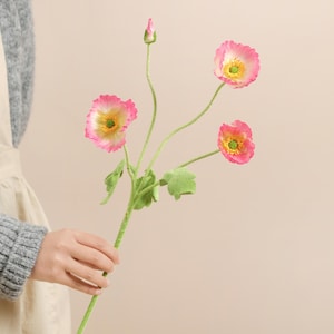 May include: A stem of artificial poppies with pink petals, yellow centres, and green stems and leaves. The flowers are in various stages of bloom, with one bud. The stem is held by a person.