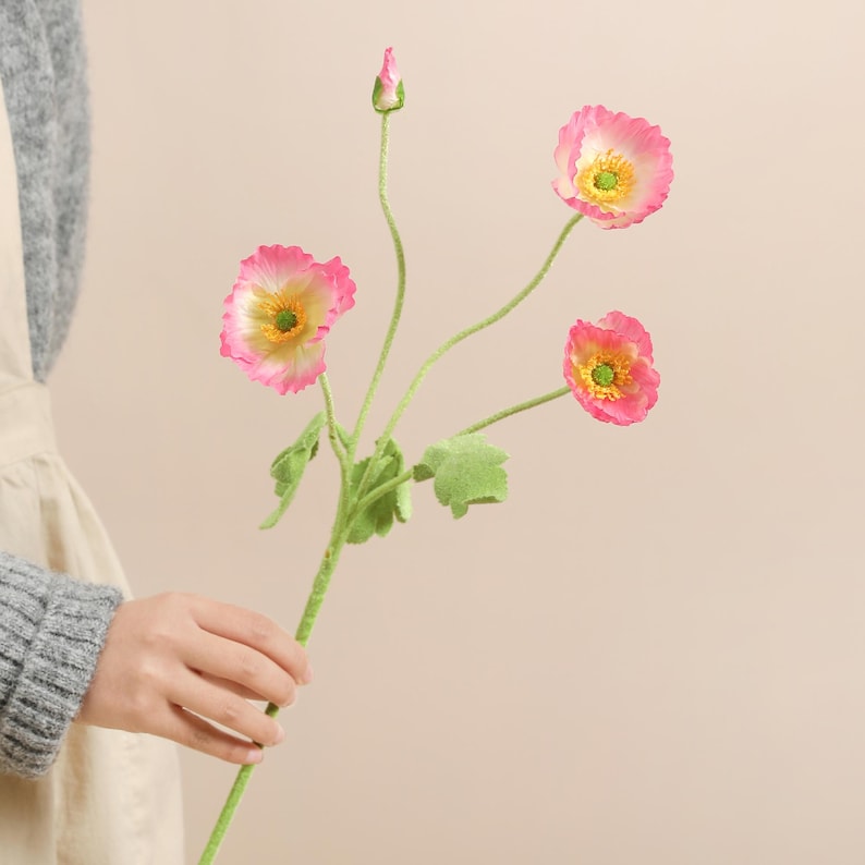 May include: A stem of artificial poppies with pink petals, yellow centres, and green stems and leaves. The flowers are in various stages of bloom, with one bud. The stem is held by a person.