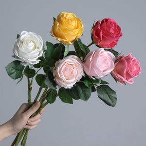 Op de afbeelding: Een boeket kunstmatige rozen in verschillende kleuren, waaronder wit, geel, roze en rood. De rozen hebben groene stelen en bladeren, vastgehouden in een hand tegen een grijze achtergrond. De bloemen staan in volle bloei.