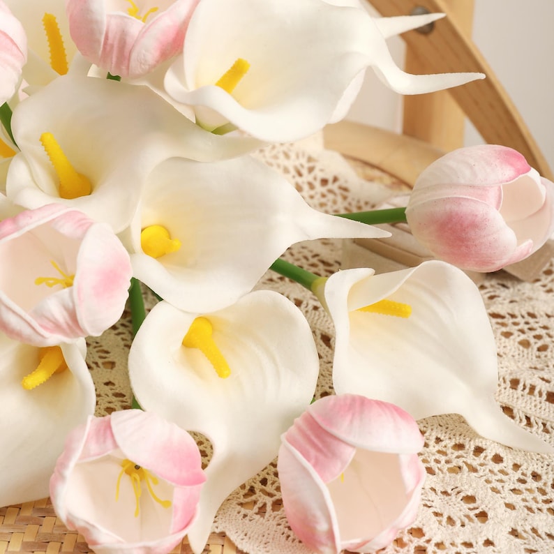 May include: A collection of artificial flowers, including white calla lilies with yellow centers and pink-tipped tulips. The flowers are arranged on a lace-like fabric, creating a soft and delicate aesthetic. The image showcases a close-up view of the blooms.