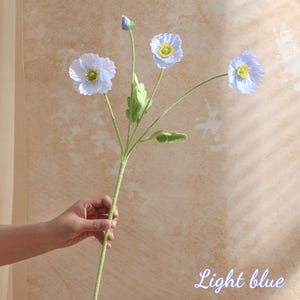 May include: A light blue artificial poppy flower stem with four blooms and a bud. The flowers have yellow centres and green leaves. The stem is green and held by a hand. The words "Light blue" are written in cursive.