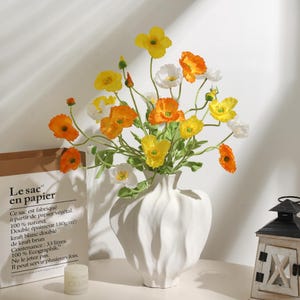 May include: A white ceramic vase holding artificial flowers in yellow, orange, and white. The vase has a textured design. A paper bag with French text and a small candle are in the background. The vase is likely around 25 cm tall.
