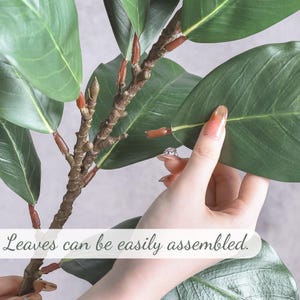 May include: An artificial plant branch featuring dark green, oval-shaped leaves attached to a textured brown stem with reddish-brown connectors. The text "Leaves can be easily assembled." is printed across the lower portion of the image.