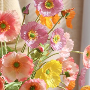 May include: A vibrant display of artificial poppies in pink, yellow, and orange hues. The flowers feature green stems and centres, with open blooms and closed buds. The arrangement is set against a soft, neutral background, creating a visually appealing display.