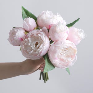 May include: A bouquet of artificial peonies featuring multiple soft pink and white layered blossoms in various stages of bloom, accompanied by several pointed green leaves and thin green stems gathered together at the base for display.