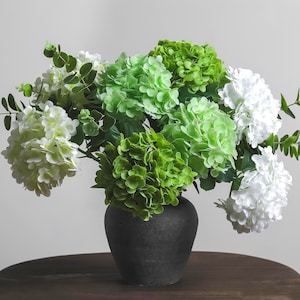May include: A bouquet of artificial hydrangea flowers in shades of white, light green, and deep green, accented with small green eucalyptus leaves. The flowers are arranged in a matte black, rounded ceramic vase with a wide base and narrow neck.