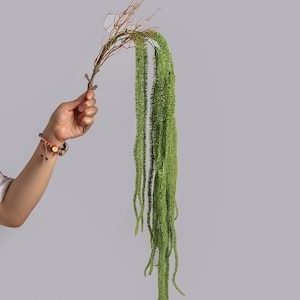 May include: A decorative artificial plant stem featuring a brown, textured branch base that transitions into multiple long, cascading, bright green foliage strands with a fine, needle-like texture that hang vertically downward.
