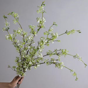 Op de afbeelding: Een boeket van kunstmatige witte bloemen met groene bladeren en stelen. De bloemen hebben kleine, delicate witte bloesems en worden in een hand gehouden tegen een grijze achtergrond. Het arrangement is een natuurlijke, elegante decoratie.