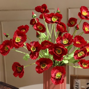 May include: A bouquet of artificial red poppy flowers with green stems and leaves arranged in a tall, vertical, light-coloured vase. Each flower features deep red petals surrounding a bright yellow and green centre stamen structure.