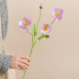 May include: A stem of artificial lavender poppies with green stems and leaves. The flowers have yellow centres and are in various stages of bloom. The background is a soft, neutral colour.
