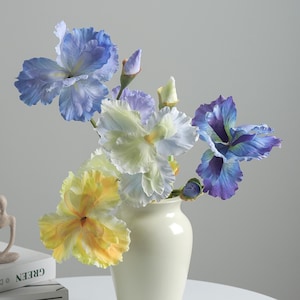 May include: A bouquet of artificial iris flowers in shades of blue, yellow, and white, featuring ruffled petals and closed buds. The flowers are arranged in a smooth, cream-colored ceramic vase with a narrow neck and a rounded base.