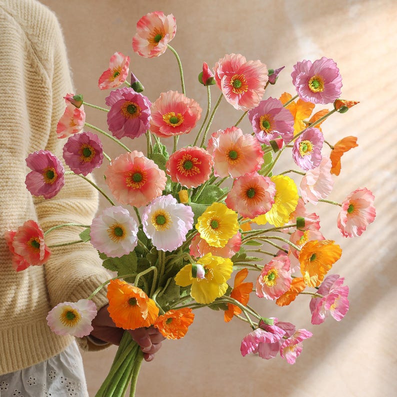 May include: A colourful bouquet of artificial poppies in shades of pink, orange, yellow, and white. The flowers have green stems and leaves, creating a cheerful arrangement. The bouquet is held in a person's hand.
