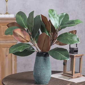 May include: A teal vase with a textured, crackled finish holds a cluster of artificial magnolia branches. The branches feature large, glossy green leaves on the top side and a matte, brownish-red underside, creating a natural contrast.
