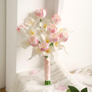 May include: A bridal bouquet featuring white calla lilies and pink tulips. The bouquet is wrapped with a light pink ribbon and green stems. Scattered pink petals add a delicate touch to the arrangement.