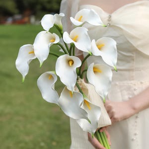 May include: A bouquet of white calla lilies with yellow centers and green stems. The flowers are held together with a cream-colored ribbon. The bouquet is a classic choice for weddings and special occasions.