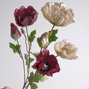 May include: A bouquet of artificial flowers featuring deep burgundy and cream-coloured poppy blossoms. The stems are thin and brown, accented with green serrated leaves and small, closed flower buds. The petals have a soft, fabric-like texture.