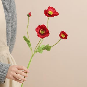 May include: A stem of artificial red poppy flowers with yellow centres and green stems and leaves. The poppy flowers are in various stages of bloom, with some fully open and others as buds.